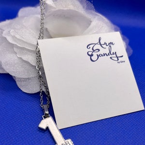 May include: A silver-toned necklace with a pendant shaped like the number one. The pendant has the Greek letters ZΦB at the base. The necklace hangs in front of a white card with the text "Arm Candy by Gina" in navy blue script. The background is a bright blue.