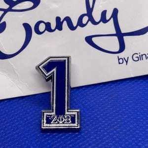 May include: A blue and silver enamel pin shaped like the number one with the letters "ZOB" inside a silver frame. The pin is on a blue background.
