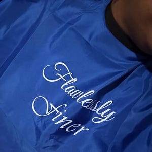 May include: A royal blue salon cape with the words "Flawlessly Finer" in elegant white script. The cape has a black neckline and is made of a smooth, shiny material, likely nylon or polyester.