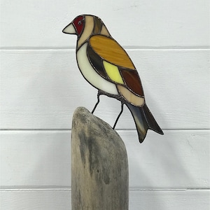 May include: A stained glass bird with brown, yellow, white and red glass pieces perched on a piece of driftwood.