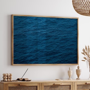 Könnte beinhalten: Ein gerahmter Druck der Meeresoberfläche, der tiefblaues Wasser mit subtilen Wellenmustern zeigt. Das Kunstwerk hängt über einem Holzschrank mit gewebten Paneelen, ergänzt durch dekorative Vasen und eine gewebte Leuchte.