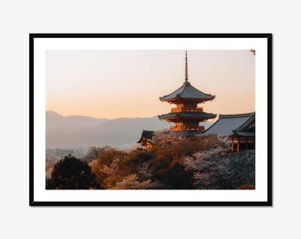 May include: A framed landscape photograph featuring a multi-tiered pagoda with dark grey roofs and orange accents, nestled among trees with light-colored blossoms. The scene is set against a backdrop of distant mountains under a soft sunset sky.