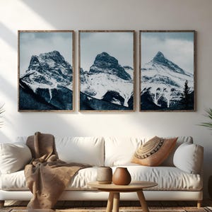 May include: Three framed prints of snow-covered mountain peaks in shades of blue and white, set against a cloudy sky. The frames are brown. The prints are displayed above a white sofa. The overall scene evokes a sense of calm.