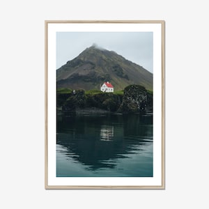 May include: A framed landscape photograph featuring a white house with a bright red roof situated on a dark, rocky cliffside. Behind the house rises a large, mist-covered mountain. The scene is reflected in the calm, dark blue water below.