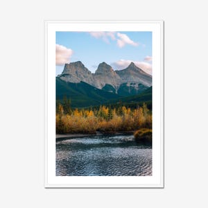 May include: A framed landscape photograph featuring a mountain range with three peaks under a blue sky with clouds. The foreground shows a body of water and trees with yellow and orange foliage.