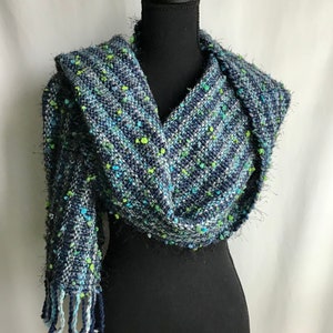 Puede incluir: Una bufanda tejida azul, verde y blanca con flecos. La bufanda está drapeada sobre un maniquí negro.