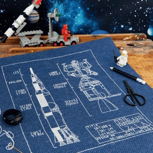 Op de afbeelding: Een blauw en wit kruissteekpatroon van een Saturnus V-raket met de titel "Saturn V Blueprint" en de tekst "Scales 1:100 1:10 Version Size" onderaan.