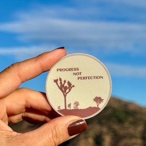 May include: A round sticker with a beige background and brown text that reads "Progress Not Perfection". The sticker features a silhouette of a Joshua Tree and other desert plants.