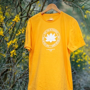 Gold t-shirt with a white round lotus that has the words, MAY YOU BE PEACEFUL-MAY YOU BE HAPPY-MAY YOU BE FREE on the front. It is hanging in front of yellow wild flowers in the Southern California desert.