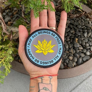May include: Round patch with a gray background, a yellow lotus flower, and the text "MAY ALL BEINGS BE HAPPY, MAY ALL BEINGS BE FREE" in a blue border. The patch is held in a hand.