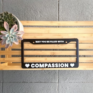 May include: Black license plate frame with white text that reads "May you be filled with COMPASSION" with a white heart on either side of the word compassion.