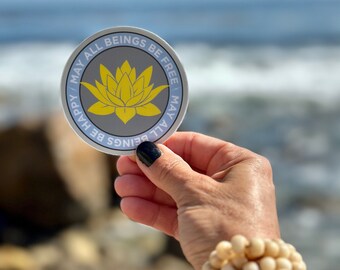 May include: A round sticker with a light blue background and a gray circle in the center. The gray circle has a yellow lotus flower in the center. The sticker has the text "May all beings be free, May all beings be happy" written around the edge.