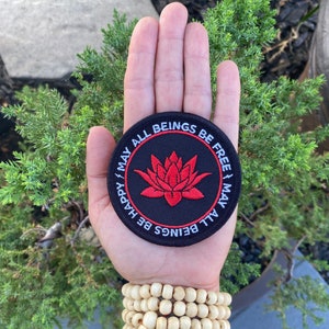 May include: A black and red embroidered patch with a white lotus flower in the center and the text "May All Beings Be Free | May All Beings Be Happy" around the edge.