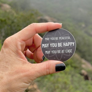 May include: A round, dark gray sticker with the text "MAY YOU BE PEACEFUL, MAY YOU BE HAPPY, MAY YOU BE AT EASE" in white. The sticker has a floral design around the edge. The sticker is held in a hand against a blurred green background.