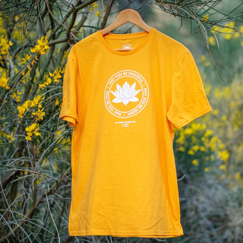 Gold t-shirt with a white round lotus that has the words, MAY YOU BE PEACEFUL-MAY YOU BE HAPPY-MAY YOU BE FREE on the front. It is hanging in front of yellow wild flowers in the Southern California desert.