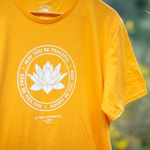 Close up of gold t-shirt with a white round lotus that has the words, MAY YOU BE PEACEFUL-MAY YOU BE HAPPY-MAY YOU BE FREE on the front. It is hanging in front of yellow wild flowers in the Southern California desert.