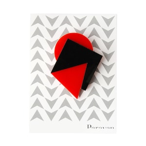 Könnte beinhalten: Rote und schwarze geometrische Brosche mit einem Chevron-Muster im Hintergrund. Die Brosche ist aus Acryl und hat ein schlichtes, modernes Design. Der Text "Paravan" ist in Schwarz am unteren Rand des Bildes gedruckt.
