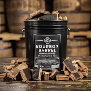 May include: A black bucket filled with bourbon barrel smoking chunks. The bucket has a white label with the text "Bourbon Barrel Smoking Chunks". Chunks are scattered around the base of the bucket. The background features wooden barrels.
