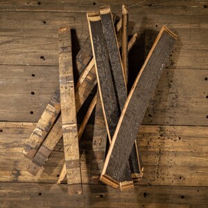 K&ouml;nnte beinhalten: Eine Sammlung von recycelten Fassdauben, die das verkohlte Innere und das verwitterte &Auml;u&szlig;ere des Holzes zeigen. Die Dauben sind auf einer rustikalen Holzoberfl&auml;che angeordnet und unterstreichen ihre einzigartigen Texturen und Farbt&ouml;ne. Das Holz hat helle und dunkle Braunt&ouml;ne.