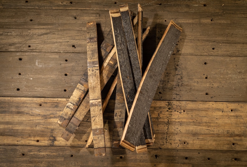 K&ouml;nnte beinhalten: Eine Sammlung von recycelten Fassdauben, die das verkohlte Innere und das verwitterte &Auml;u&szlig;ere des Holzes zeigen. Die Dauben sind auf einer rustikalen Holzoberfl&auml;che angeordnet und unterstreichen ihre einzigartigen Texturen und Farbt&ouml;ne. Das Holz hat helle und dunkle Braunt&ouml;ne.