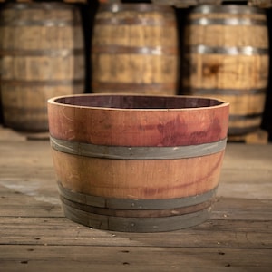 May include: A small, open-top wooden barrel with a reddish-brown interior and exterior. The barrel is held together with several metal bands. The background features several larger wooden barrels.