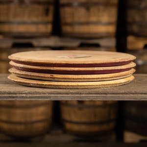 May include: A stack of four round, wooden barrel lids with a reddish-brown band around the edge. The lids are light brown with visible wood grain and sit on a dark wooden shelf. The background features more wooden barrels.