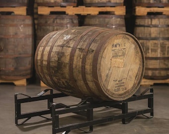 May include: A weathered wooden barrel with metal bands, resting on a black metal stand. The barrel has a faded label with text. Other barrels are stacked in the background.