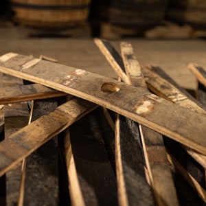 K&ouml;nnte beinhalten: Nahaufnahme von verwitterten Holzfassdauben, einige mit einem kleinen Loch, verstreut auf einer Holzoberfl&auml;che. Die Dauben sind in verschiedenen Braun- und Graut&ouml;nen gehalten, mit sichtbarer Holzmaserung und Textur. Der Hintergrund zeigt verschwommene Holzf&auml;sser.
