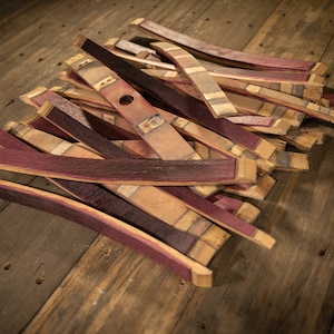 May include: A collection of reclaimed wooden barrel staves, showcasing a mix of natural wood tones and deep purple hues. Some staves have a hole and markings, suggesting their origin. The pieces are arranged on a rustic wooden surface.
