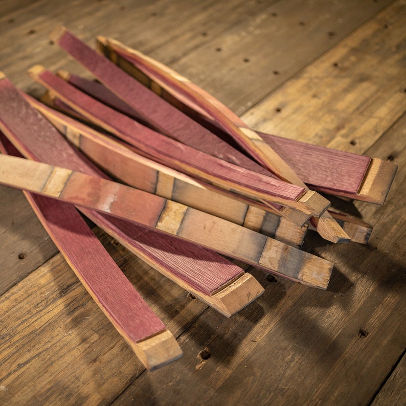May include: A collection of reclaimed wood barrel staves, featuring a rich burgundy interior and natural wood exterior. The staves are cut and shaped, with visible grain and variations in color, resting on a rustic wooden surface.