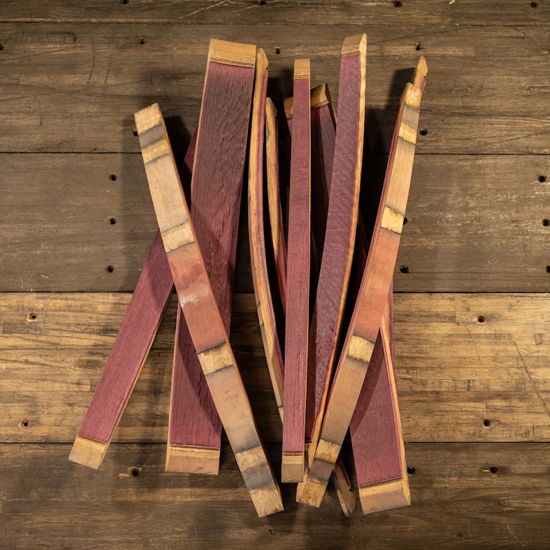 May include: A collection of reclaimed, curved wooden barrel staves. The staves are a deep purple-red color, with lighter wood accents at the ends and along the sides. The wood has a rustic, weathered appearance.