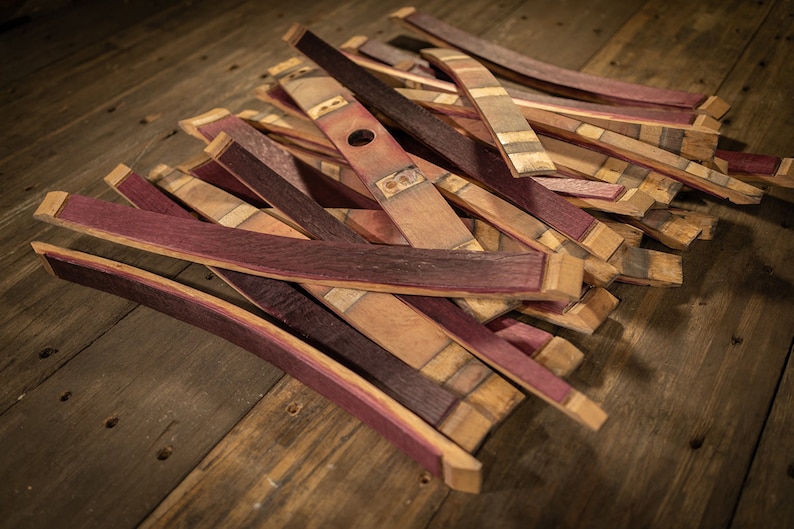 May include: A collection of reclaimed wood barrel staves, featuring a mix of colors including deep red, natural wood tones, and hints of purple. The staves are cut and shaped, with some showing metal hardware.
