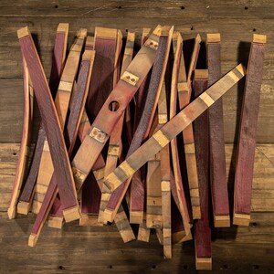 May include: A collection of reclaimed wine barrel staves, showcasing a mix of reddish-purple and natural wood tones. Some staves have metal bands and a hole, suggesting their origin. The staves are arranged on a rustic wooden surface.