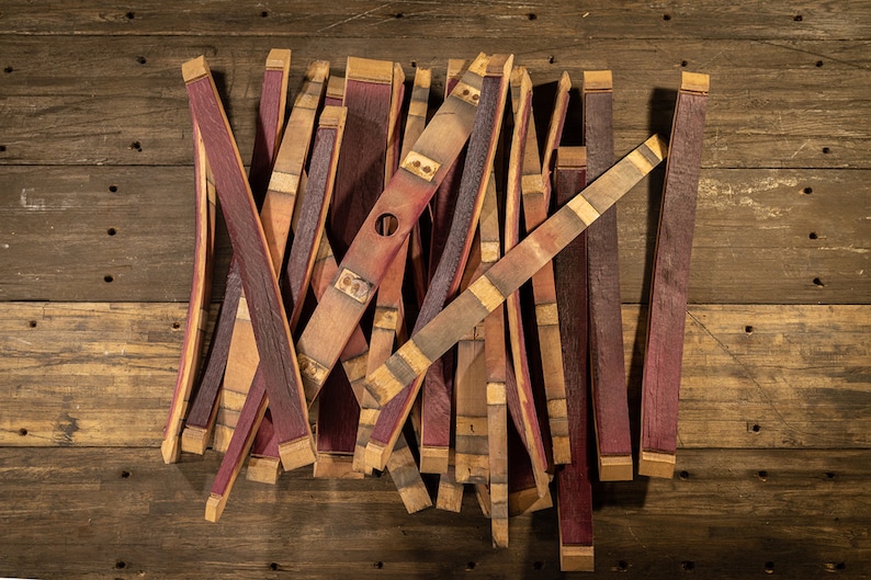 May include: A collection of reclaimed wine barrel staves, showcasing a mix of reddish-purple and natural wood tones. Some staves have metal bands and a hole, suggesting their origin. The staves are arranged on a rustic wooden surface.