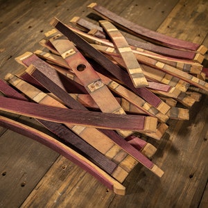 May include: A pile of reclaimed wood barrel staves, featuring a mix of natural wood tones and deep burgundy hues. Some staves have a hole and metal hardware. The staves are arranged on a rustic wooden surface.