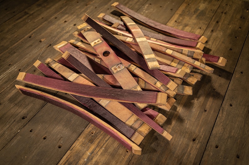 May include: A pile of reclaimed wood barrel staves, featuring a mix of natural wood tones and deep burgundy hues. Some staves have a hole and metal hardware. The staves are arranged on a rustic wooden surface.