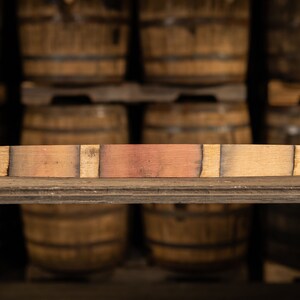 May include: A rectangular piece of reclaimed wood, likely from a barrel, with varying shades of brown and reddish-brown. The wood is placed horizontally on a wooden shelf, with a blurred background of stacked wooden barrels.