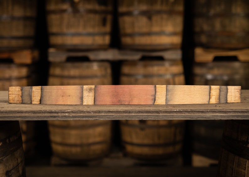 May include: A rectangular piece of reclaimed wood, likely from a barrel, with varying shades of brown and reddish-brown. The wood is placed horizontally on a wooden shelf, with a blurred background of stacked wooden barrels.