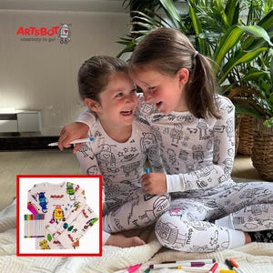 May include: Two young girls wearing white pajamas with black robot designs, laughing and holding markers. The pajamas are part of a coloring set, as shown in the inset image, which includes a set of colored markers. The logo "ArtsBot" is visible in the top left corner.