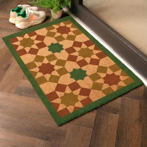 May include: A rectangular doormat featuring a geometric star design in brown, green, and tan hues. The mat has a green border and is positioned near a door. Trainers and a potted plant are also visible.
