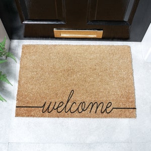 Könnte beinhalten: Eine braune Kokosmatte mit dem Wort "welcome" in schwarzer Kursivschrift.