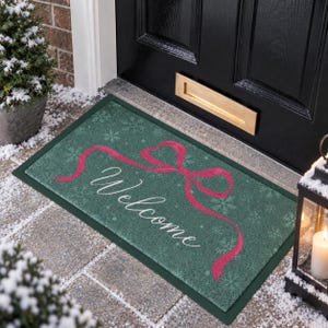 May include: A rectangular doormat with a dark green background featuring a white snowflake pattern, a large pink ribbon graphic, and the word Welcome written in white cursive script across the centre of the mat surface.