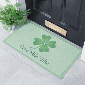 Può includere: Uno zerbino rettangolare verde chiaro con una grafica di un quadrifoglio verde scuro e il testo Céad Míle Fáilte stampato in un carattere corsivo verde scuro. Il tappetino presenta un bordo solido e una superficie testurizzata su piastrelle.