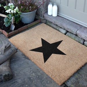 May include: A rectangular coir doormat with a large black star design. The mat is made from natural fibres and is placed on a stone surface. The background includes plants in a metal bucket and a pair of wellington boots.
