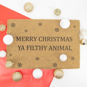 May include: A brown coir doormat with the text "Merry Christmas Ya Filthy Animal" in black lettering. The doormat is surrounded by gold and white Christmas ornaments.