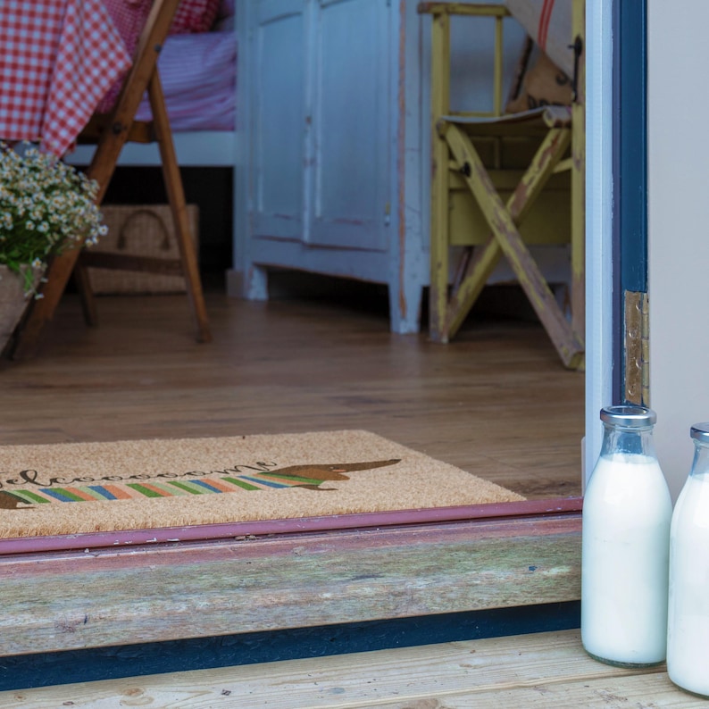 May include: A coir doormat with a dachshund design and the word "welcome" in cursive. Two glass milk bottles with silver caps sit on a wooden surface. The scene suggests a rustic, country-style setting.