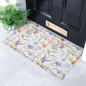 May include: Rectangular doormat with a floral pattern featuring purple, yellow, pink, and blue flowers with green leaves on a cream background. The mat is placed in front of a black door with a gold letter slot.