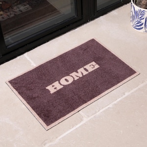 May include: A rectangular doormat in a deep purple colour, with the word "HOME" in a bold, off-white font. The mat has a lighter border and is placed on a light-coloured stone surface, suitable for an entryway.