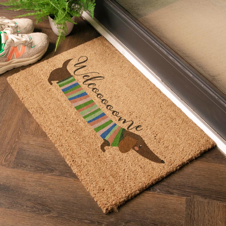 May include: A rectangular doormat made of natural fibers. It features a brown dachshund wearing a striped sweater in shades of blue, green, and pink. The word "Welcome" is written above the dog in a script font. The mat is placed near a doorway.