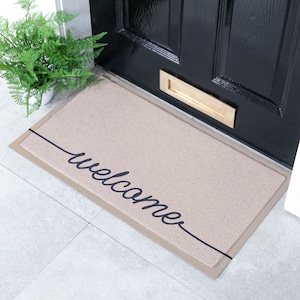 May include: A beige rectangular doormat with the word "welcome" in black script. A black line runs diagonally across the mat. The mat is positioned in front of a black door with a gold letterbox.